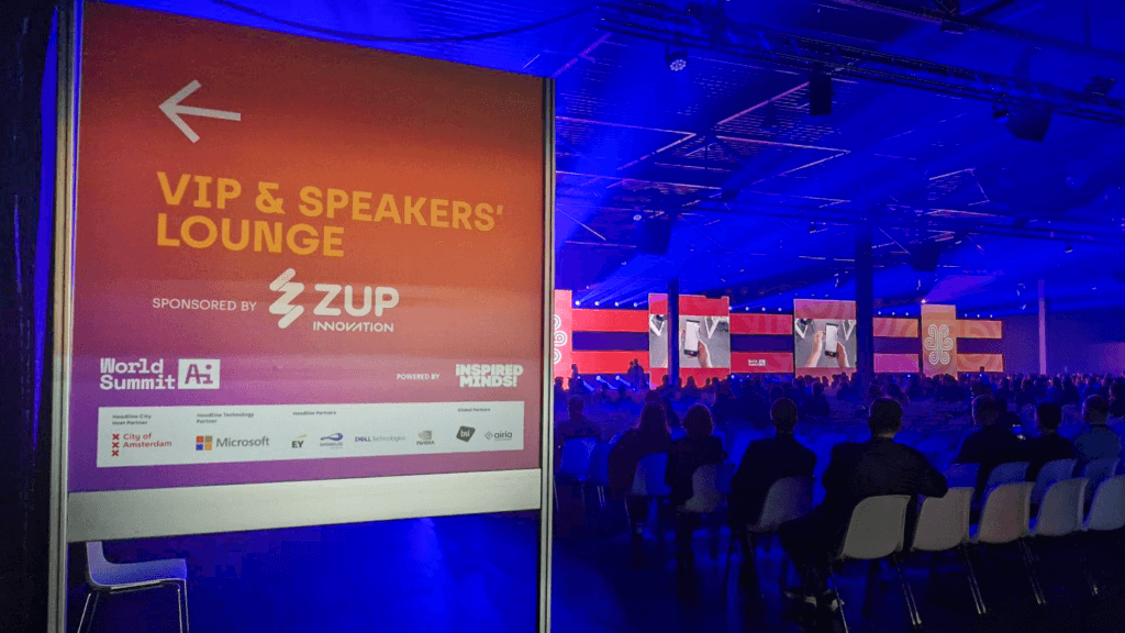 Um grande espaço onde as pessoas estão sentadas participando do evento World Summit AI 2024, assistindo a uma palestra ao fundo. Em destaque, podemos ver um banner indicando a localização do lounge para participantes VIPs e palestrantes, com a indicação do patrocínio da Zup Innovation. 
