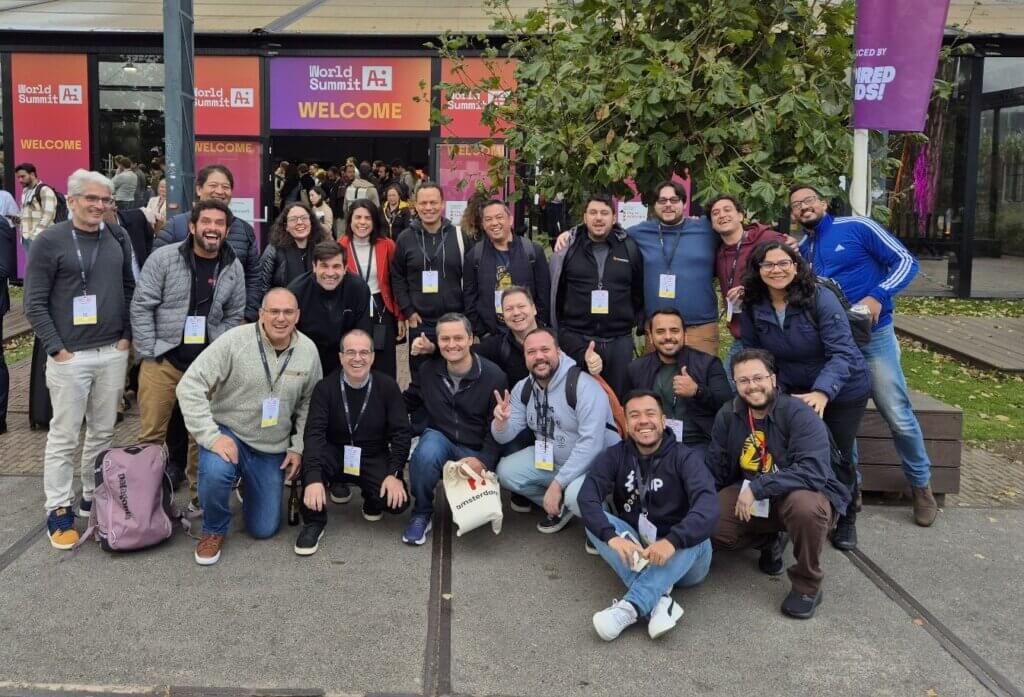  Um grupo diverso de pessoas sorrindo e posando juntos para uma fotografia ao ar livre, atrás do grupo é possível ler “World Summit AI Welcome”.