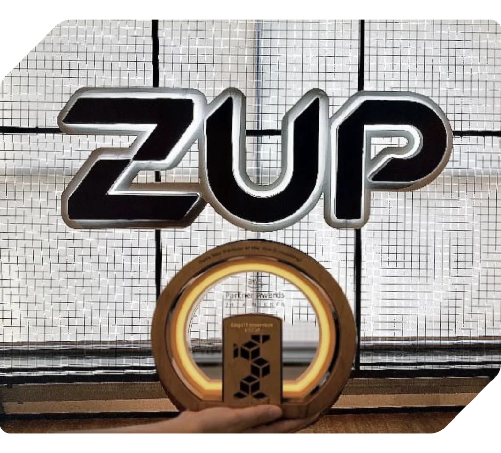Imagem institucional com logotipo da Zup em destaque sobre fundo industrial, enquanto uma mão de pele clara segura um objeto circular dourado, representando tecnologia e inovação.