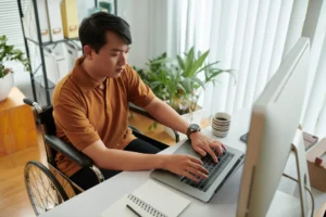 Fotografia de um profissional de tecnologia asiático em uma cadeira de rodas, trabalhando concentrado em seu notebook sobre uma mesa branca de escritório. A imagem serve de ilustração para o tema IA generativa na modernização de legado: 6 práticas essenciais. No ambiente moderno e iluminado, veem-se um monitor de computador, uma caneca de café, plantas decorativas e uma estante com arquivos organizadores ao fundo.