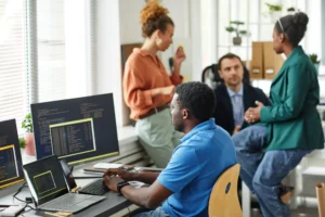 Imagem capa do conteúdo sobre Modernização de sistemas legados para engenharia sênior, onde há uma fotografia em um escritório moderno e bem iluminado. Em primeiro plano, um desenvolvedor de software negro, vestindo uma camisa polo azul, trabalha concentrado em sua mesa de costas para a câmera. À sua frente, há dois monitores e um notebook abertos exibindo linhas de código em telas escuras. Ao fundo, três colegas de trabalho conversam: uma mulher com blusa laranja e cabelos cacheados presos, um homem de paletó escuro e uma mulher negra vestindo blazer verde, sentada de forma descontraída na borda de uma mesa. O ambiente corporativo conta com grandes janelas com persianas, plantas decorativas e prateleiras com organizadores de arquivos.