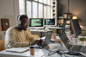 Fotografia de um engenheiro de software negro focado, com fones de ouvido e óculos, digitando em seu laptop em um escritório moderno de plano aberto. Vários monitores ao fundo exibem códigos de programação, simbolizando o complexo gerenciamento de tráfego de APIs que ele executa. Na mesa, um notebook de anotações e um café descartável. Esta cena exemplifica o trabalho de otimização de engenharia detalhado no caso de sucesso: Veja como reduzimos em 80% o tráfego de APIs para uma multinacional de telefonia utilizando o Amazon Bedrock. Acesse o caso de sucesso.