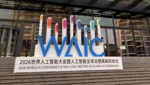 Uma fotografia da entrada de um moderno edifício de convenções em Xangai, com um grande letreiro tridimensional da 'WAIC' (World AI Conference). A estrutura tridimensional apresenta as letras 'WAIC' em azul grande e tridimensional, com colunas de barras de progresso coloridas (gradientes de amarelo, rosa, roxo e azul-claro) acima de cada letra. Abaixo da estrutura, na base branca, o texto completo lê-se: '2026世界人工智能大会暨人工智能全球治理高级别会议' e '2026 WORLD AI CONFERENCE & HIGH-LEVEL MEETING ON GLOBAL AI GOVERNANCE'. O letreiro está posicionado sobre degraus de pedra em frente à fachada de vidro do edifício.