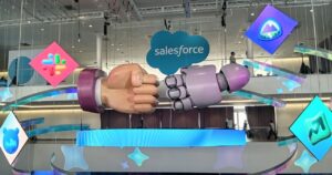 Imagem capa do conteúdo sobre Agentforce e a Era Agêntica: o papel da engenharia de software no futuro da IA. Uma instalação de arte suspensa em um grande átrio de conferência moderno exibe um "fist bump" simbólico e colaborativo entre um grande punho humano e um grande punho de robô metálico. Acima deles, um grande logotipo da nuvem da Salesforce com a palavra "salesforce" em letras brancas está centralizado. O punho do robô é feito de metal polido e o punho humano é realisticamente esculpido. Esta interação simboliza a colaboração entre a inteligência artificial e a humanidade. Ao redor da instalação principal, formas geométricas facetadas e logotipos menores abstratos de produtos da Salesforce (incluindo um urso, um gráfico de crescimento e um ícone de estrela de mídia social) também estão suspensos. O fundo mostra a escala do átrio com passarelas e algumas pessoas borradas em um nível superior, sob uma iluminação interna limpa e cabos finos de suspensão visíveis.