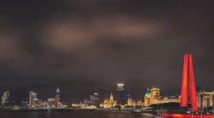 Imagem capa do conteúdo sobre ecossistemas de ia agenda estrategica. Onde há um cenário noturno panorâmico de Xangai, na China, capturado ao longo do rio Huangpu. O famoso horizonte da Bund é iluminado, mostrando uma fileira de edifícios históricos e modernos com arquitetura variada e luzes douradas, refletindo suavemente na água. O Monumento aos Heróis do Povo de Xangai, com sua distinta estrutura de colunas vermelhas em forma de trípode, está proeminentemente iluminado em vermelho brilhante no primeiro plano à direita. O céu está escuro e encoberto, criando uma atmosfera dramática sobre a paisagem urbana.