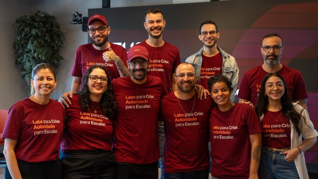 Uma fotografia de plano médio em ambiente interno mostra um grupo de dez pessoas, composto por homens e mulheres de diversas idades, posando juntos e sorridentes para a câmera, dispostos em duas fileiras (uma à frente e outra atrás).A grande maioria das pessoas veste uma camiseta de cor vinho escuro, na qual se lê no peito o texto em letras laranjas e brancas: "Labs para Criar. Autoridade para Escalar." Alguns homens usam bonés também na cor vinho, sendo que um deles (atrás, à esquerda) faz um sinal de "joinha" com a mão direita e tem tatuagens visíveis no braço. Um dos homens na fileira de trás veste uma jaqueta jeans aberta sobre a camiseta.As pessoas estão posicionadas de forma descontraída, algumas com os braços ao redor dos ombros dos colegas, transmitindo um clima de união, colaboração e espírito de equipe. O grupo exibe expressões alegres e amigáveis.Ao fundo, vislumbra-se um espaço de escritório ou evento, com uma parede escura, uma planta ornamental à esquerda e iluminação interna natural. A composição foca no grupo, que ocupa a maior parte do enquadramento, celebrando um momento coletivo.