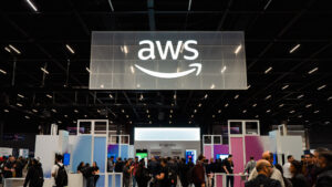 Fotografia de um grande pavilhão de exposições movimentado durante o AWS Summit 2026, com muitas pessoas caminhando entre estandes coloridos e iluminados. No alto, pendurado no teto escuro e cheio de estruturas de iluminação, destaca-se um enorme painel translúcido e iluminado com o logotipo da AWS (Amazon Web Services) em branco. Abaixo, o público transita pelo espaço do evento, onde é possível ver estandes com painéis nas cores azul, branco e rosa, telas digitais e identificações como "IA Agêntica". Diversos participantes usam mochilas e crachás, caracterizando o ambiente corporativo e tecnológico do AWS Summit 2026. Espectadores e expositores conversam e exploram as atrações do evento