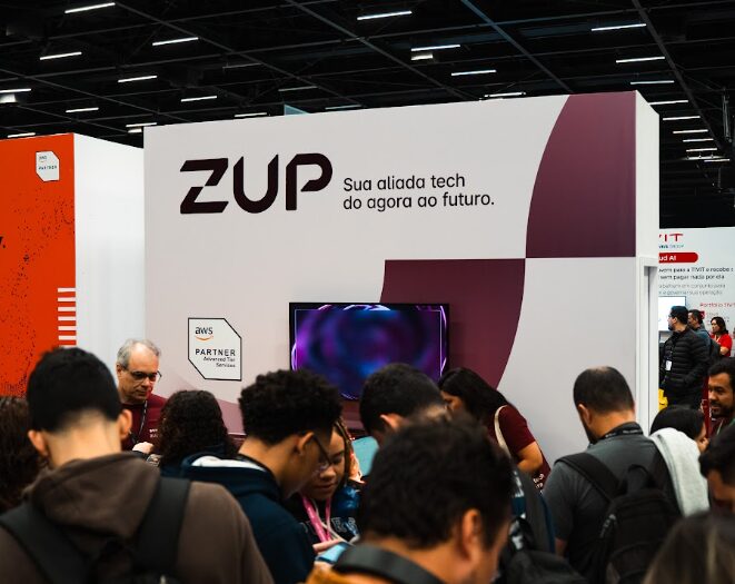 Fotografia do stand da Zup no AWS Summit 2026. O expositor branco apresenta o logótipo da Zup em destaque, acompanhado do slogan "Sua aliada tech do agora ao futuro" e do selo "AWS Partner". Em primeiro plano, um grupo de participantes do evento encontra-se reunido em frente ao stand, onde é visível uma tela.